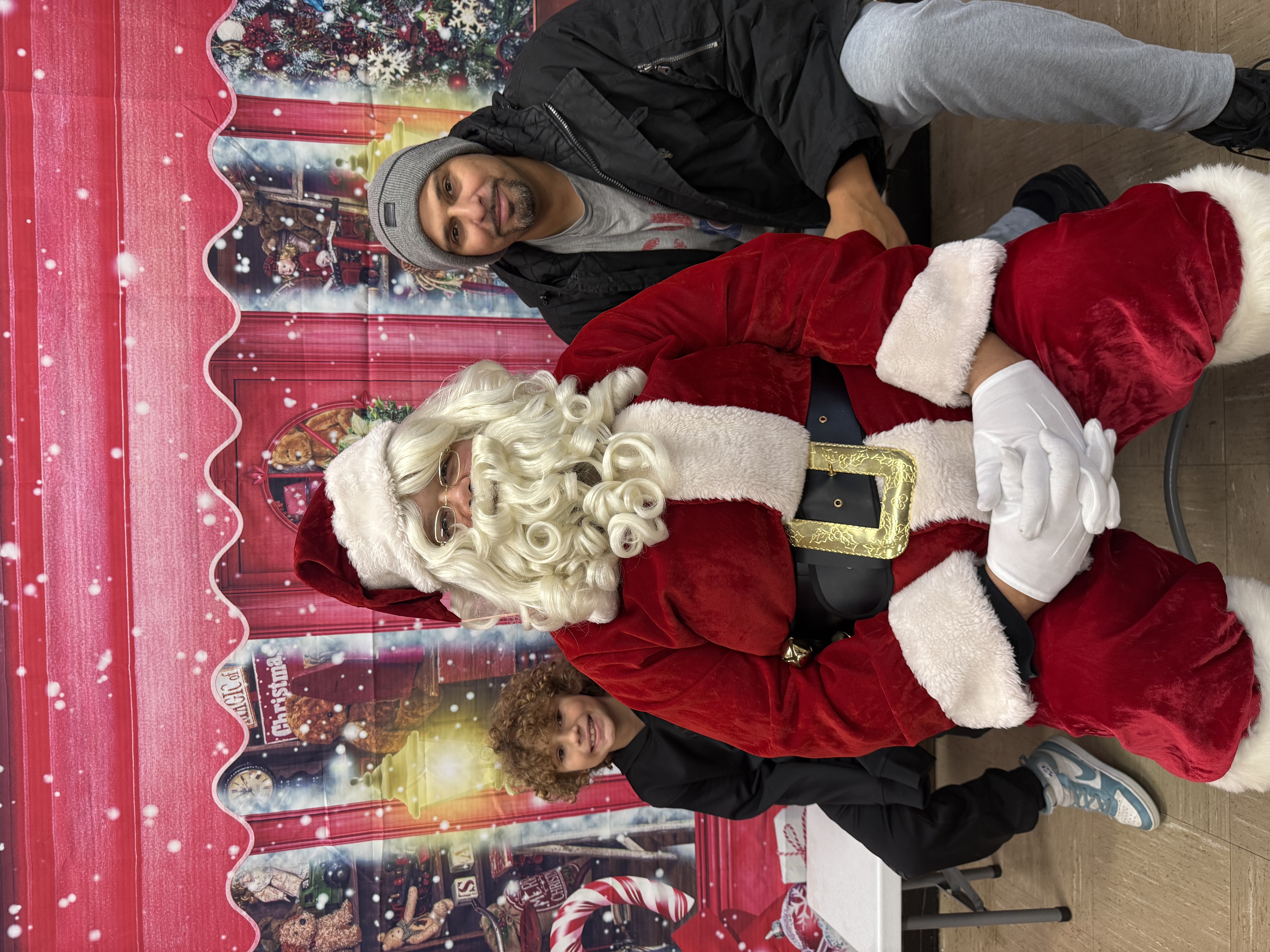 Photograph of Santa with two people, in front of a holiday backdrop.
